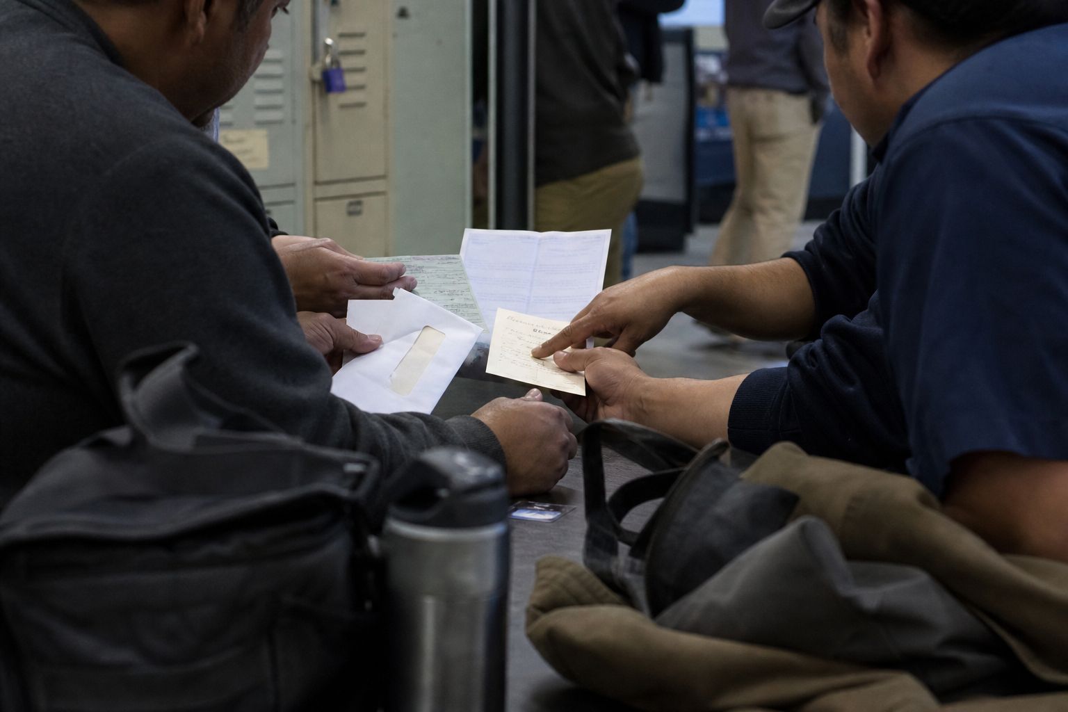 Two coworkers at the lockers compare their pay stubs side by side.